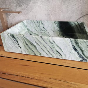 May include: A rectangular, green and white marble sink with a natural, flowing pattern. The sink is set on a wooden surface, with a gold-colored faucet in the background. The sink's design is modern and minimalist.
