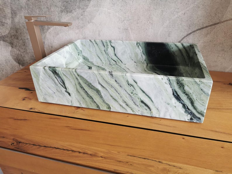 May include: A rectangular, green and white marble sink with a natural, flowing pattern. The sink is set on a wooden surface, with a gold-colored faucet in the background. The sink's design is modern and minimalist.