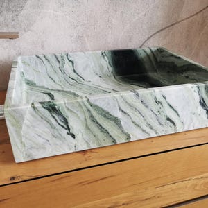 May include: A rectangular, marble sink with a natural green and white veined pattern. The sink is set on a wooden surface, showcasing its angular design and polished finish. The sink's interior is a deep, dark green.