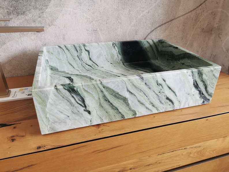May include: A rectangular, marble sink with a natural green and white veined pattern. The sink is set on a wooden surface, showcasing its angular design and polished finish. The sink's interior is a deep, dark green.