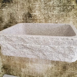 Könnte beinhalten: Ein weißes Steinkesselbecken mit einer rauen, strukturierten Außenseite. Das Becken ist rechteckig und hat einen leicht abgerundeten Rand.