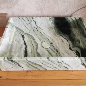 May include: Rectangular bathroom sink with a natural marble pattern in shades of green, black, and white. The sink has a central drain and is set on a wooden surface. A gold-colored faucet is visible on the left.