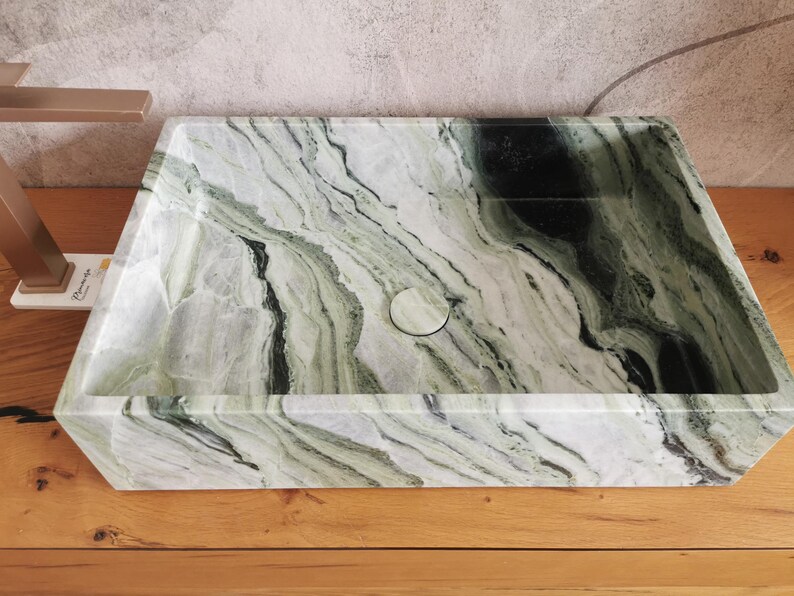 May include: Rectangular bathroom sink with a natural marble pattern in shades of green, black, and white. The sink has a central drain and is set on a wooden surface. A gold-colored faucet is visible on the left.