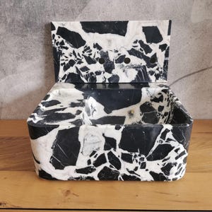 May include: A black and white marble container with a hinged lid. The container has a rectangular shape with rounded corners and a textured surface. The lid has two circular holes. The marble pattern is a mix of black and white.