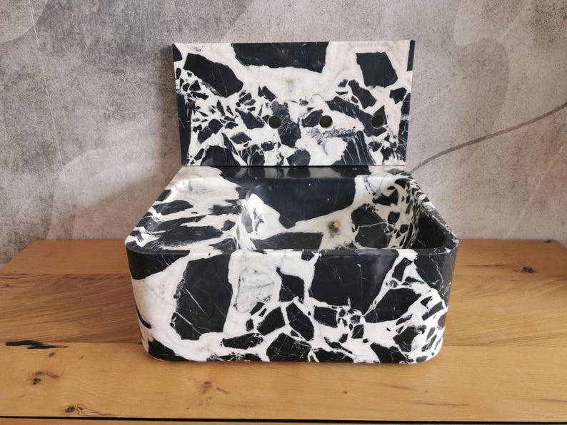 May include: A black and white marble container with a hinged lid. The container has a rectangular shape with rounded corners and a textured surface. The lid has two circular holes. The marble pattern is a mix of black and white.