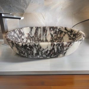 May include: A white marble bathroom sink with black and purple veining. The sink is oval shaped and sits on a white countertop.