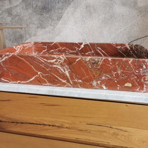 May include: A rectangular red marble sink with white veining. The sink is sitting on a white countertop and a wooden cabinet.