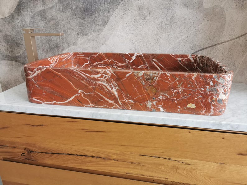 May include: A rectangular red marble sink with white veining. The sink is sitting on a white countertop and a wooden cabinet.