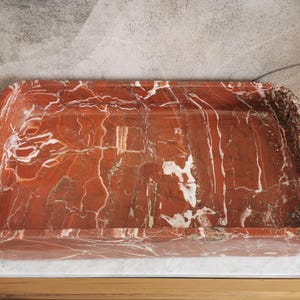 May include: A rectangular red marble sink with white veining. The sink is made of natural stone and has a smooth, polished finish.