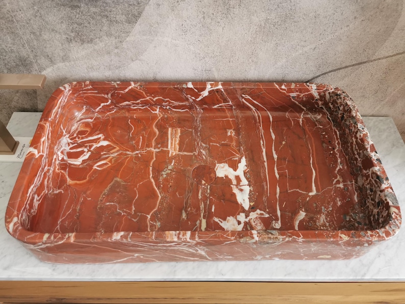 May include: A rectangular red marble sink with white veining. The sink is made of natural stone and has a smooth, polished finish.