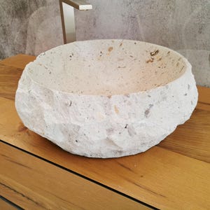 Lavabo rotondo in marmo beige visto lateralmente con lavorazione esterna grezza e tonalità crema