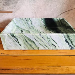 May include: Rectangular marble sink with a green and white veined pattern. A gold-colored faucet is positioned to the left. The sink is set on a wooden surface. The sink's dimensions are approximately 24 inches long, 16 inches wide, and 6 inches deep.