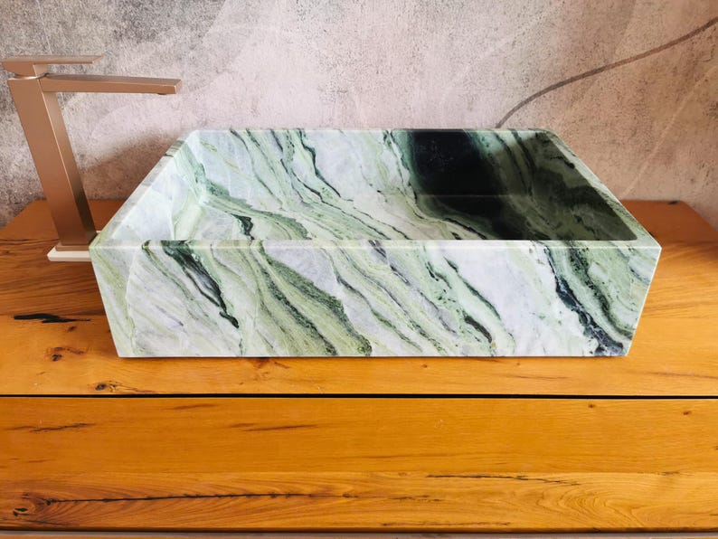 May include: Rectangular marble sink with a green and white veined pattern. A gold-colored faucet is positioned to the left. The sink is set on a wooden surface. The sink's dimensions are approximately 24 inches long, 16 inches wide, and 6 inches deep.