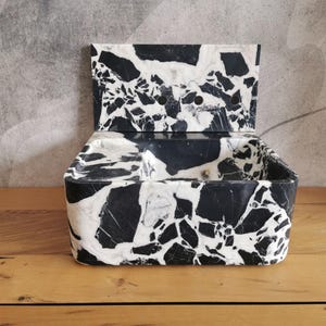 May include: A black and white marble-patterned box with a hinged lid. The box has a rectangular shape with rounded corners and a smooth, polished finish. The lid is open, revealing the interior of the box. The box is sitting on a wooden surface.