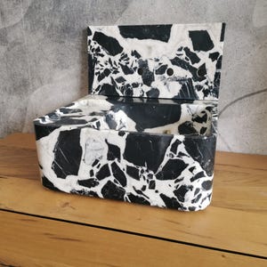 May include: A rectangular box with a hinged lid, crafted from a black and white terrazzo-style material. The box is open, revealing its interior. The design features a striking contrast of black and white, creating a modern aesthetic.