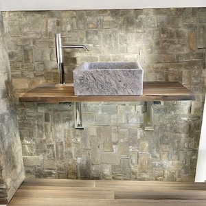 Peut inclure: Un évier en pierre grise avec un robinet chromé monté sur un comptoir en bois. Le comptoir est soutenu par des supports métalliques et le mur derrière l'évier est fait de carreaux de pierre empilés.
