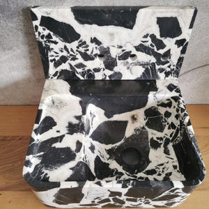 May include: A black and white marble sink with a unique, angular design. The sink features a rectangular basin with a curved front and a raised, angled back. The marble has a striking pattern of black and white veining.