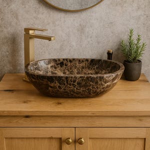 May include: Oval-shaped marble bathroom sink with brown and beige veining, set on a light wood cabinet. A gold faucet and round mirror are visible. A small potted plant and bottle are on the countertop.