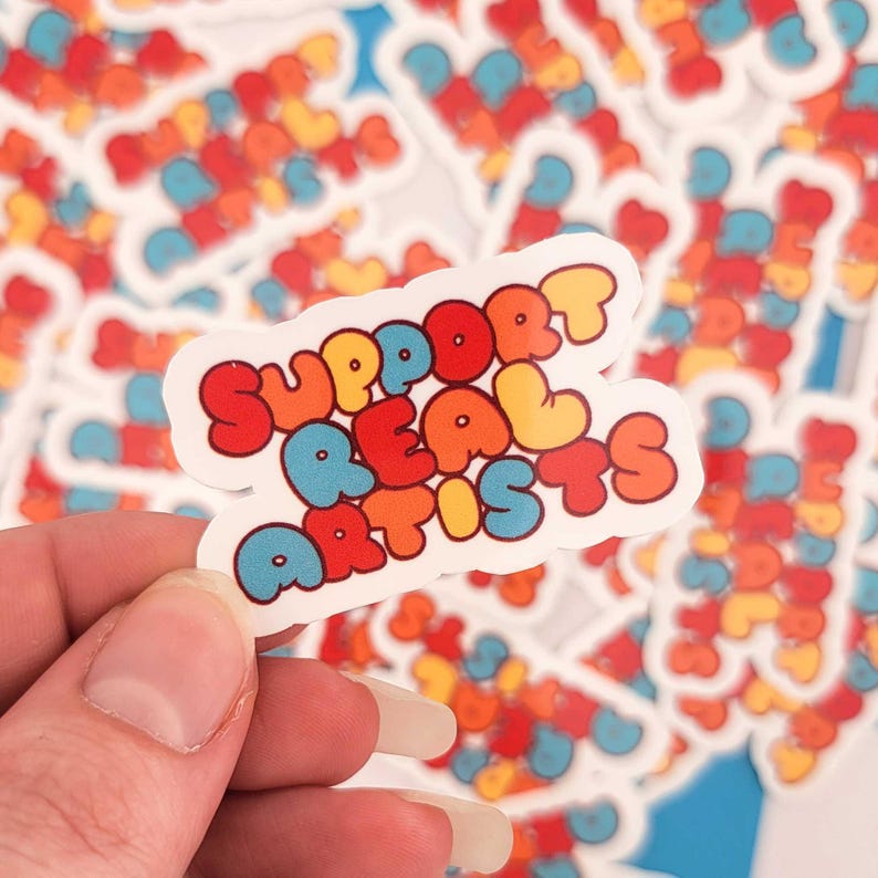 Puede incluir: Una pegatina colorida con el texto "SUPPORT REAL ARTISTS" en letras de burbujas. Las letras son rojas, naranjas, amarillas y azules. La pegatina se sostiene en una mano, con otras pegatinas similares en el fondo.