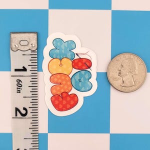 Puede incluir: Una pegatina troquelada con la palabra "Stitch" en letras de burbujas. La pegatina mide aproximadamente 3,8 cm de ancho. Las letras son en tonos de azul, naranja y rojo, con patrones de corazones y estrellas. Una moneda est&aacute; al lado de la pegatina para comparar el tama&ntilde;o.
