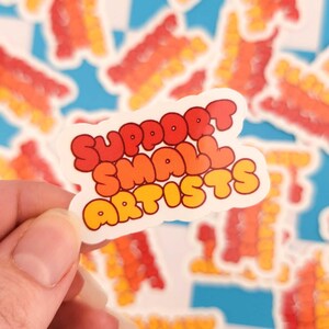 Puede incluir: Una pegatina blanca con el texto "SUPPORT SMALL ARTISTS" en letras de burbujas rojas, naranjas y amarillas. La pegatina est&aacute; sujeta por una mano, con otras pegatinas similares en el fondo. Las pegatinas tienen un dise&ntilde;o retro.