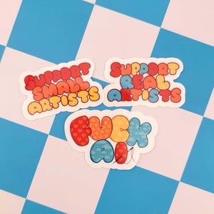 Puede incluir: Tres pegatinas coloridas con letras de burbujas sobre un fondo a cuadros. Las pegatinas dicen "SUPPORT SMALL ARTISTS", "SUPPORT REAL ARTISTS" y "FUCK AI". Las letras son rojas, naranjas, amarillas, azules y blancas.