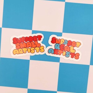 Puede incluir: Dos pegatinas con el texto "SUPPORT SMALL ARTISTS" y "SUPPORT REAL ARTISTS" en fuente de burbujas. El texto es rojo, naranja, amarillo y azul. Las pegatinas est&aacute;n sobre un fondo a cuadros azul y blanco.