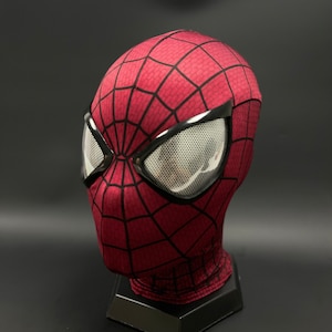 May include: A red and black Spider-Man mask with a mesh eye covering. The mask is on a black stand.