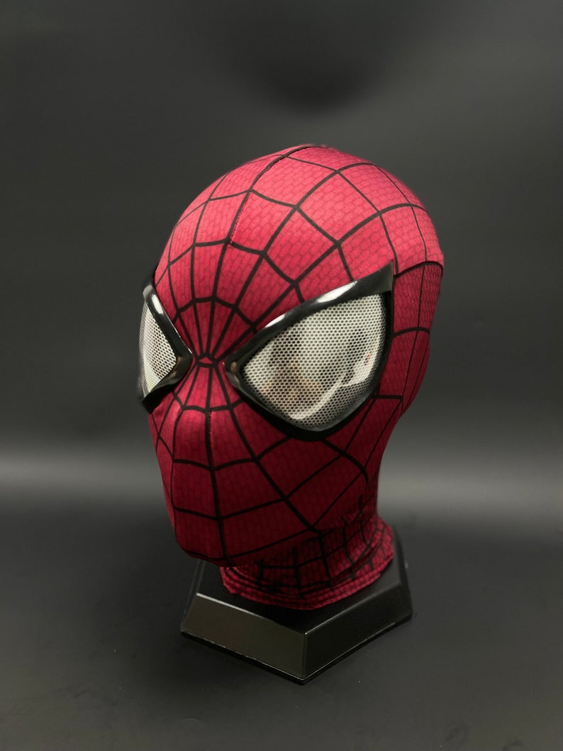 May include: A red and black Spider-Man mask with a mesh eye covering. The mask is on a black stand.
