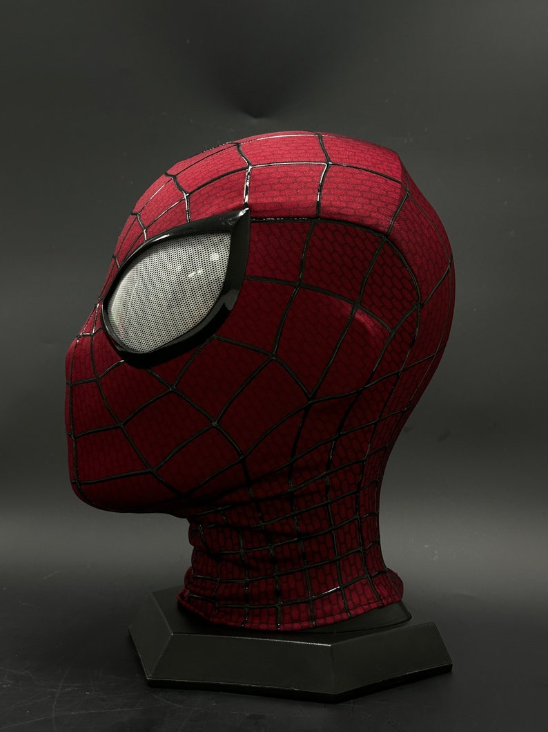May include: A red and black Spider-Man mask with a black mesh eye covering. The mask is on a black stand.