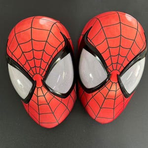 May include: Two red Spider-Man masks with black webbing and black eye frames. The eye openings are white with a mesh texture. The masks are a vibrant red color and are likely made of a flexible material.
