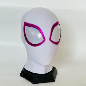 Könnte beinhalten: Eine weiße Spider-Gwen-Maske mit rosa Augenrahmen und Augenabdeckungen aus Mesh. Die Maske steht auf einem schwarzen Sockel, und der Kopf hat eine strukturierte Oberfläche.