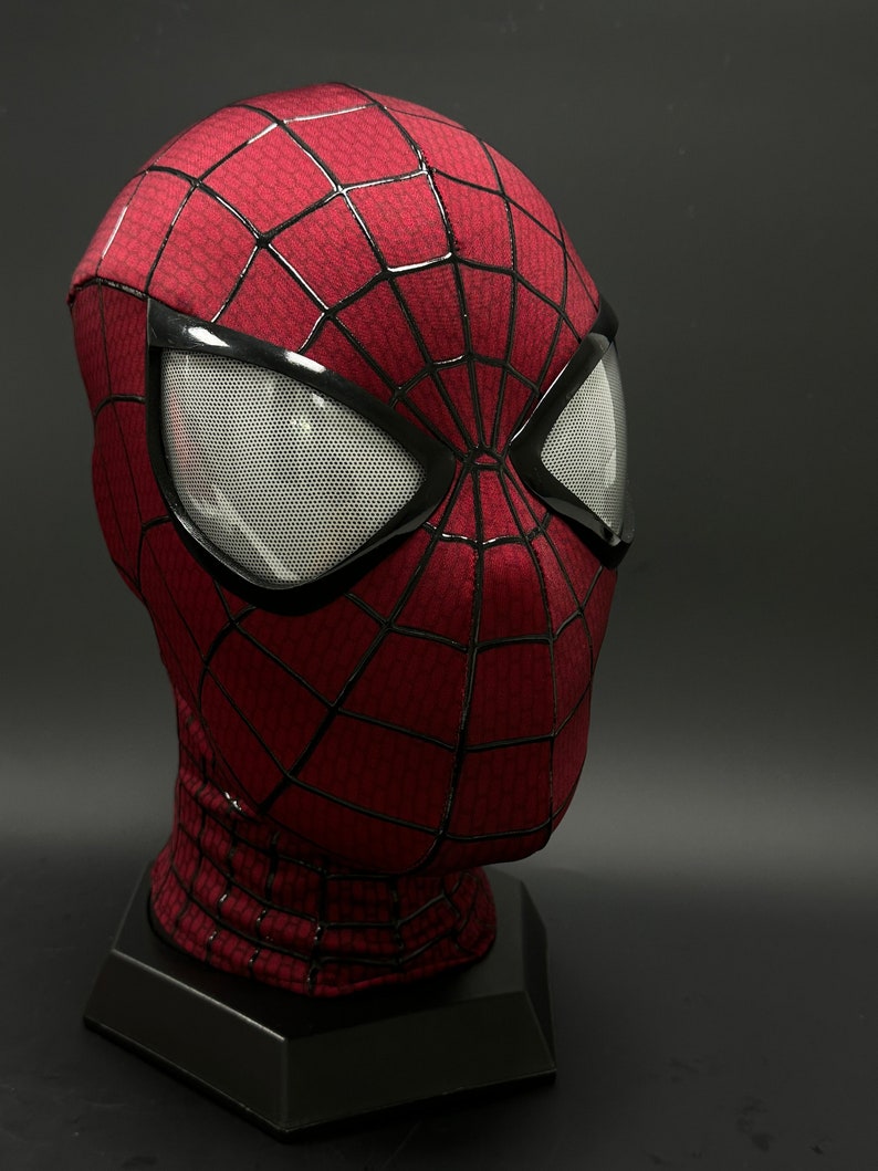 May include: A red and black Spider-Man mask with a mesh eye covering. The mask is on a black stand.