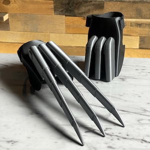 May include: A pair of black and silver claw-like costume accessories. The claws are made of a hard plastic and are attached to a black fabric base. The claws are sharp and pointed, and the base is designed to fit around the hand.