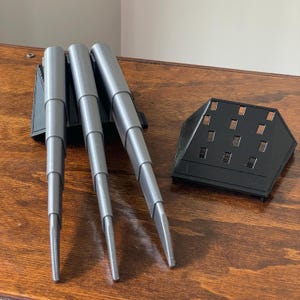 Peut inclure: Trois griffes grises articulées avec une base noire et un support noir séparé. Les griffes sont faites d'un matériau lisse d'aspect métallique et sont conçues pour être portées. Le support comporte de multiples découpes rectangulaires.
