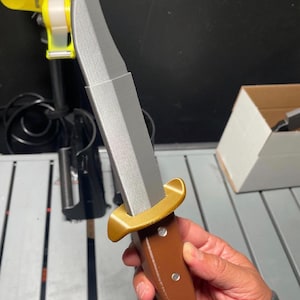 May include: A large knife with a silver blade and a brown handle. The handle has a gold-coloured guard. The knife is held in a person's hand. The background is blurred, but includes a yellow clamp and a box.