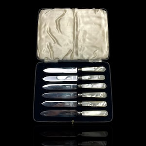 May include: A set of six silver butter knives with mother-of-pearl handles, presented in a black velvet-lined case with a satin interior. The knives are arranged neatly, reflecting in the glossy surface below.