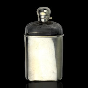 May include: A silver-colored flask with a clear, patterned glass top and a rounded, clear glass stopper. The flask has a cylindrical body and a reflective surface, set against a black background.