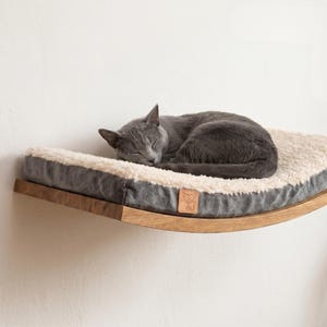 May include: A curved wooden wall-mounted cat shelf featuring a rectangular cushion with a cream-colored faux fur top and dark gray sides. A small tan leather tag with a cat face icon is attached to the front edge of the cushion.