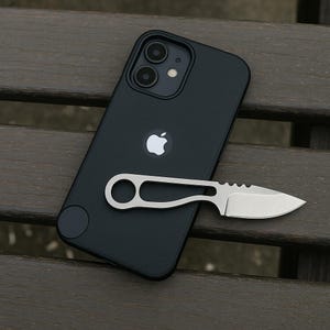 以下が含まれることがあります： デュアルレンズカメラシステムと中央にシルバーのAppleロゴを備えたマットブラックのスマートフォン。指を通すための円形の穴が開いたハンドルを持つ、小型のシルバーカラーの金属製固定刃ナイフが、デバイスの背面に水平に置かれています。
