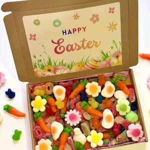 May include: An open cardboard box filled with colorful Easter-themed candies. The box features a decorative lid with the words "Happy Easter" and floral and egg designs. The candies include carrot, flower, and egg shapes, along with other assorted sweets.