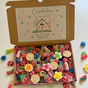 Pode incluir: Uma caixa de presente de cartão cheia de doces coloridos, incluindo ursinhos de goma, flores e anéis. A tampa da caixa apresenta um cartão impresso com o texto "Parabéns pela sua nova casa" e uma ilustração de uma casa.