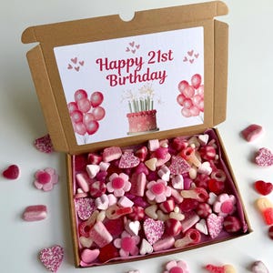 May include: A brown cardboard gift box filled with a variety of pink and red sweets. The box lid displays a card with the text "Happy 21st Birthday", balloons, and a cake illustration. Sweets include hearts, flowers, and gummy shapes.