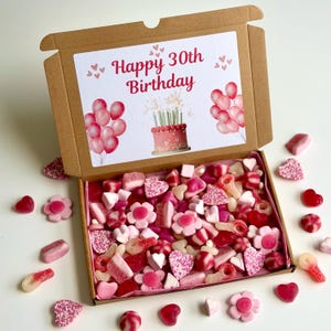 Könnte beinhalten: Eine braune Geschenkbox aus Pappe, gefüllt mit rosa und roten Süßigkeiten. Der Deckel der Box zeigt "Happy 30th Birthday", Ballons und einen Kuchen. Verschiedene Süßigkeiten sind um die Box herum verstreut.