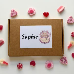 May include: A brown cardboard box with a white label that says "Sophie" and a "Happy Birthday" cake illustration. The box is surrounded by various candies, including heart-shaped and flower-shaped gummies, and pink and white sweets.