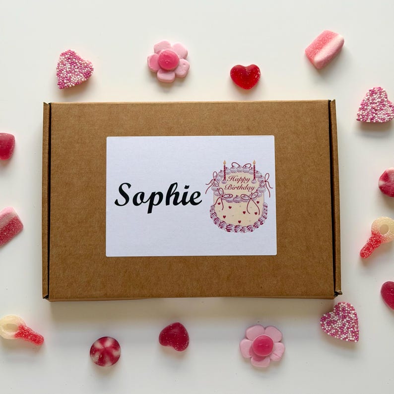 May include: A brown cardboard box with a white label that says "Sophie" and a "Happy Birthday" cake illustration. The box is surrounded by various candies, including heart-shaped and flower-shaped gummies, and pink and white sweets.