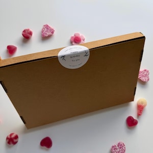 May include: A brown cardboard box with a white sticker that reads "Happy Birthday To You" surrounded by floral designs. Scattered around the box are pink candies in various shapes, including hearts and flowers.