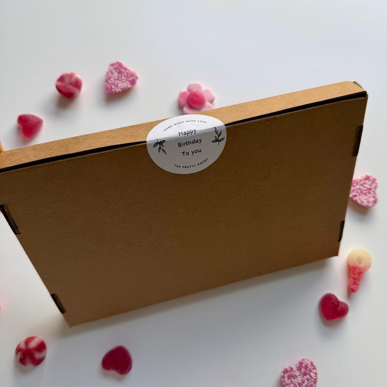 May include: A brown cardboard box with a white sticker that reads "Happy Birthday To You" surrounded by floral designs. Scattered around the box are pink candies in various shapes, including hearts and flowers.