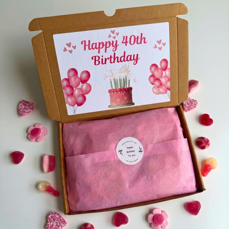 May include: A brown cardboard box with a white card that reads "Happy 40th Birthday" with pink balloons and a cake illustration. The box contains pink tissue paper and is surrounded by pink candies.