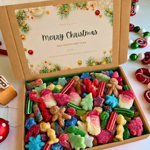 May include: A cardboard gift box filled with assorted Christmas-themed gummy candies, including gingerbread people, Santa Claus figures, and stars. The lid features a printed greeting that reads Merry Christmas and Happy New Year.